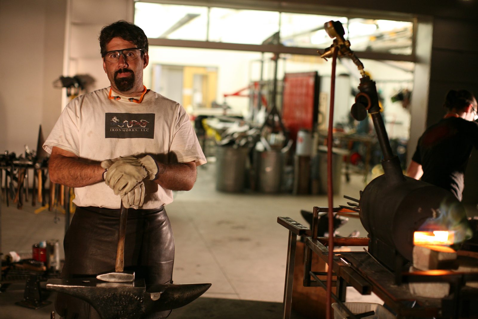 Inside the Dragonforge shop - Michael at the anvil, the coal hearth glowing on the right, tool wall behind.