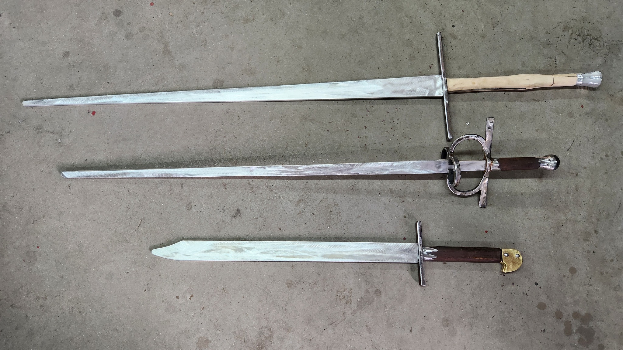 Three hand-forged swords laid out on concrete.